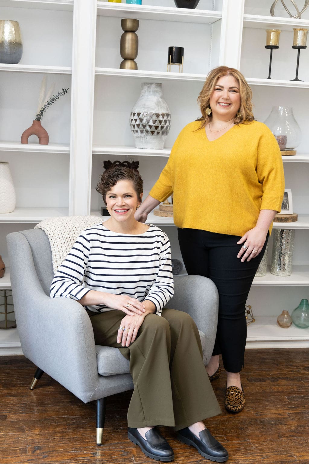 Therapist mentor standing and somatic coaching provider sitting in front of a bookshelf and smiling at the camera.