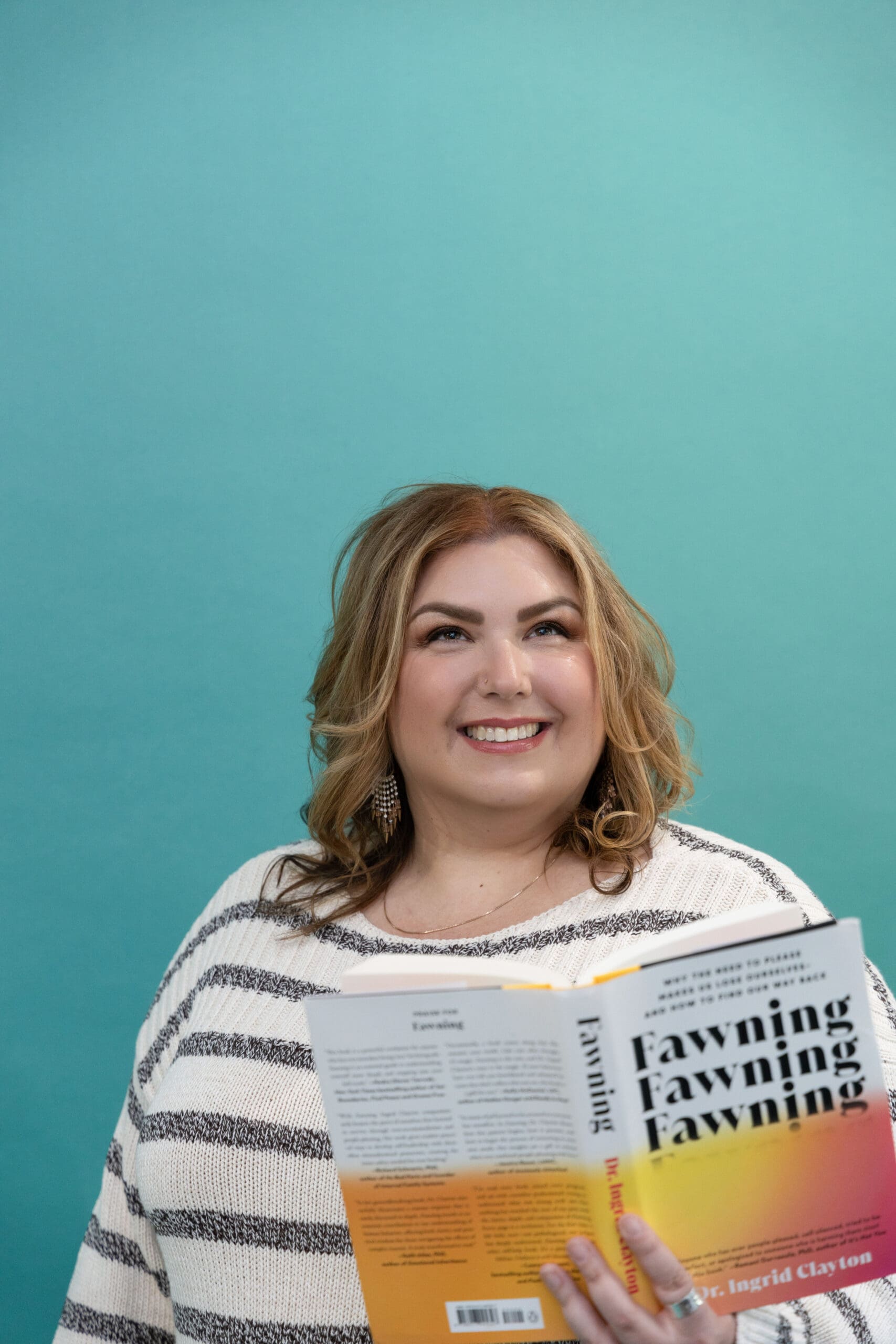 Somatic healing practitioner holding a book titled “Fawning” and smiling off into the distance