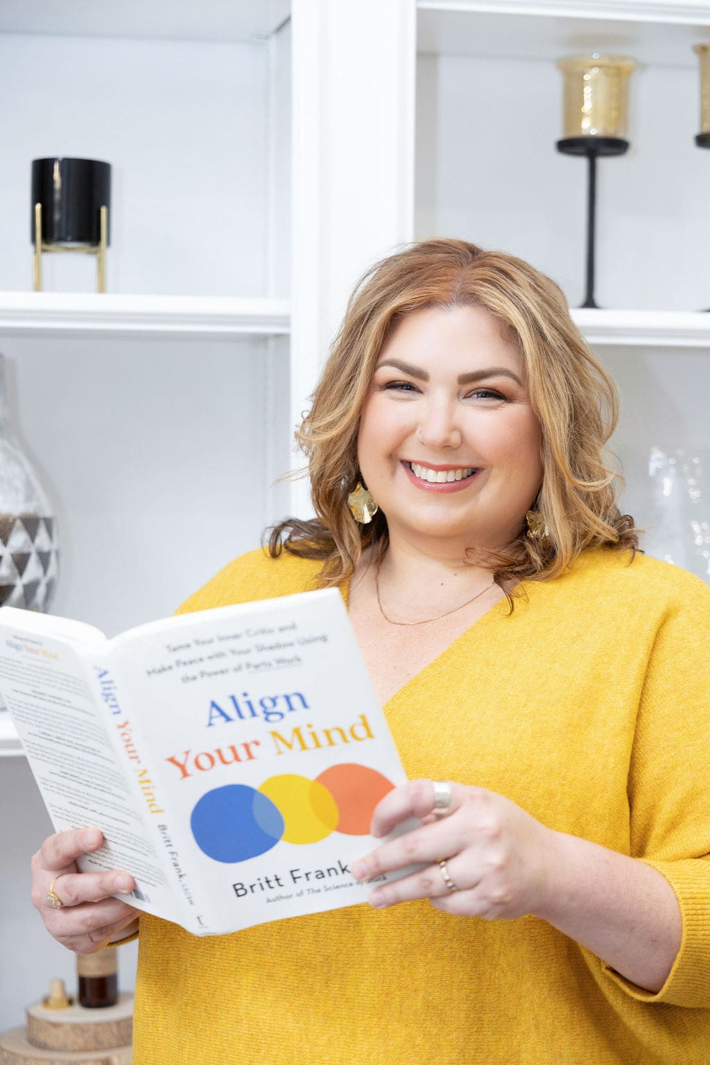 Therapist mentor and somatic experiencing practitioner standing in front of a book shelf holding a book and reading.