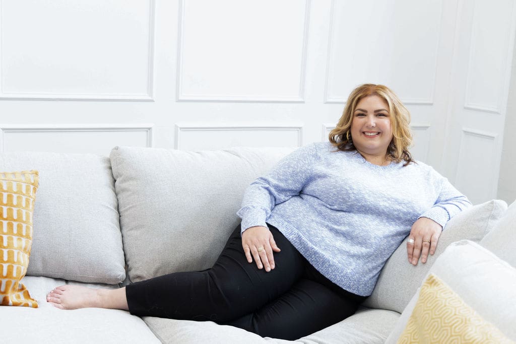 Therapist mentor and somatic experiencing practitioner sitting on a couch in jeans and a blue sweater, smiling at the camera.
