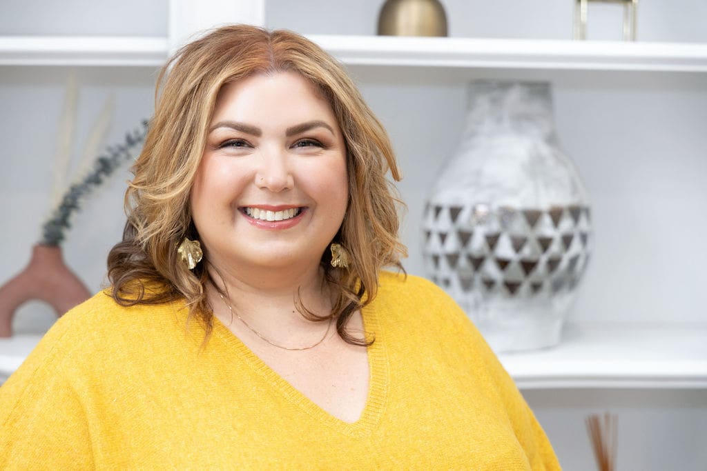 The Empowered Therapist, somatic experiencing practitioner, standing in yellow shirt in front of a bookshelf smiling