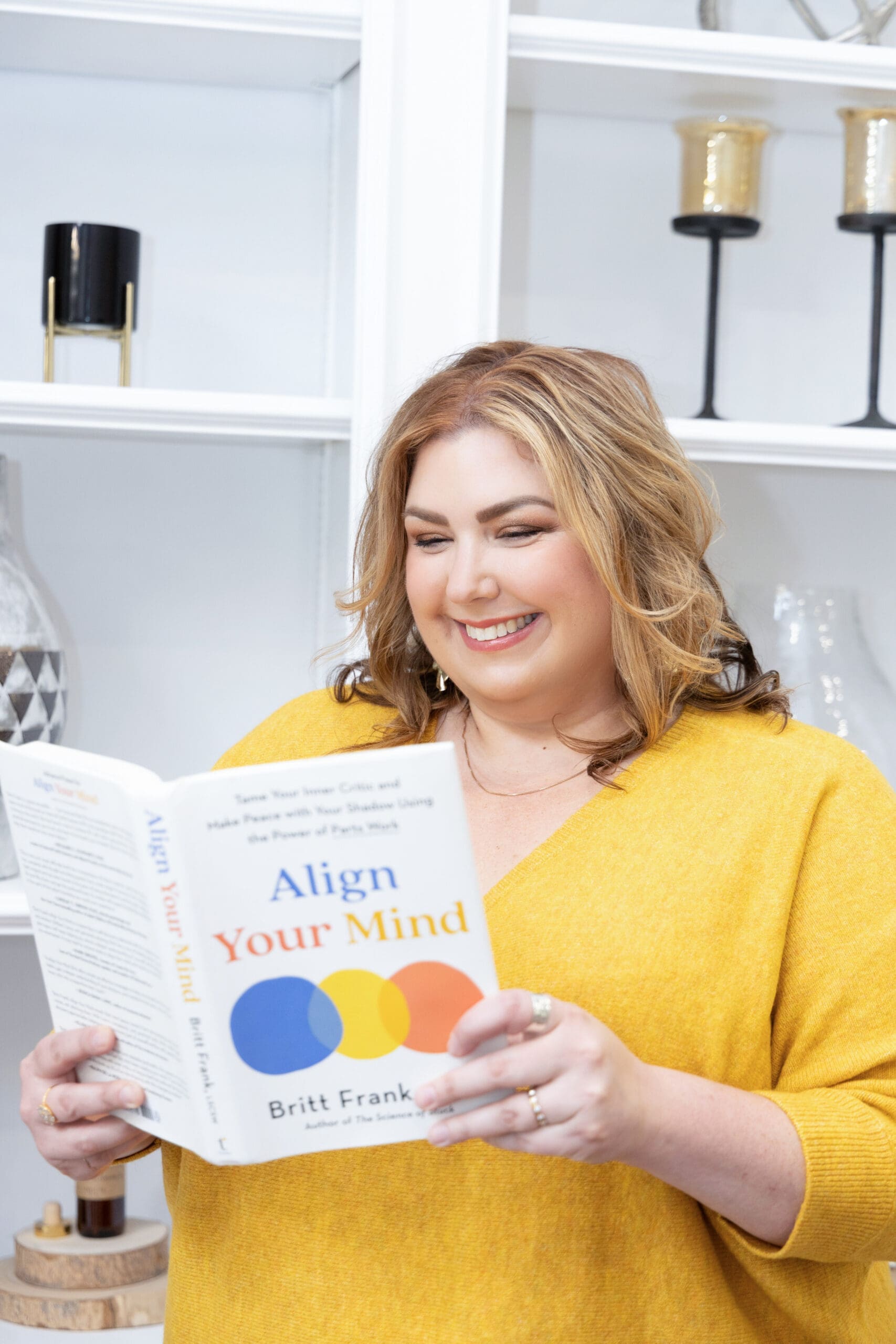The empowered therapist and somatic experiencing practitioner holding and reading a book in front of a bookcase, wearing a yellow shirt.