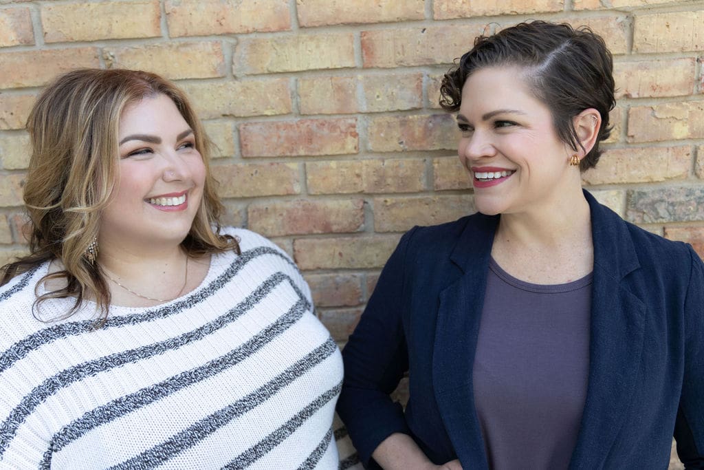 Therapist mentor talking to somatic experiencing practitioner while standing in front of a brick wall and smiling at one another.