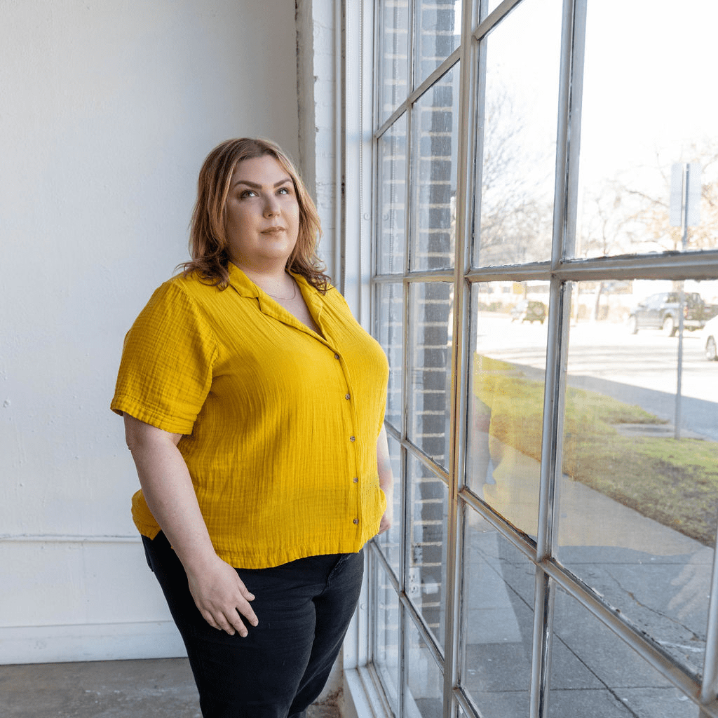 The Empowered Therapist standing in front of a wall of windows in black jeans and a yellow shirt, gazing into the distance.