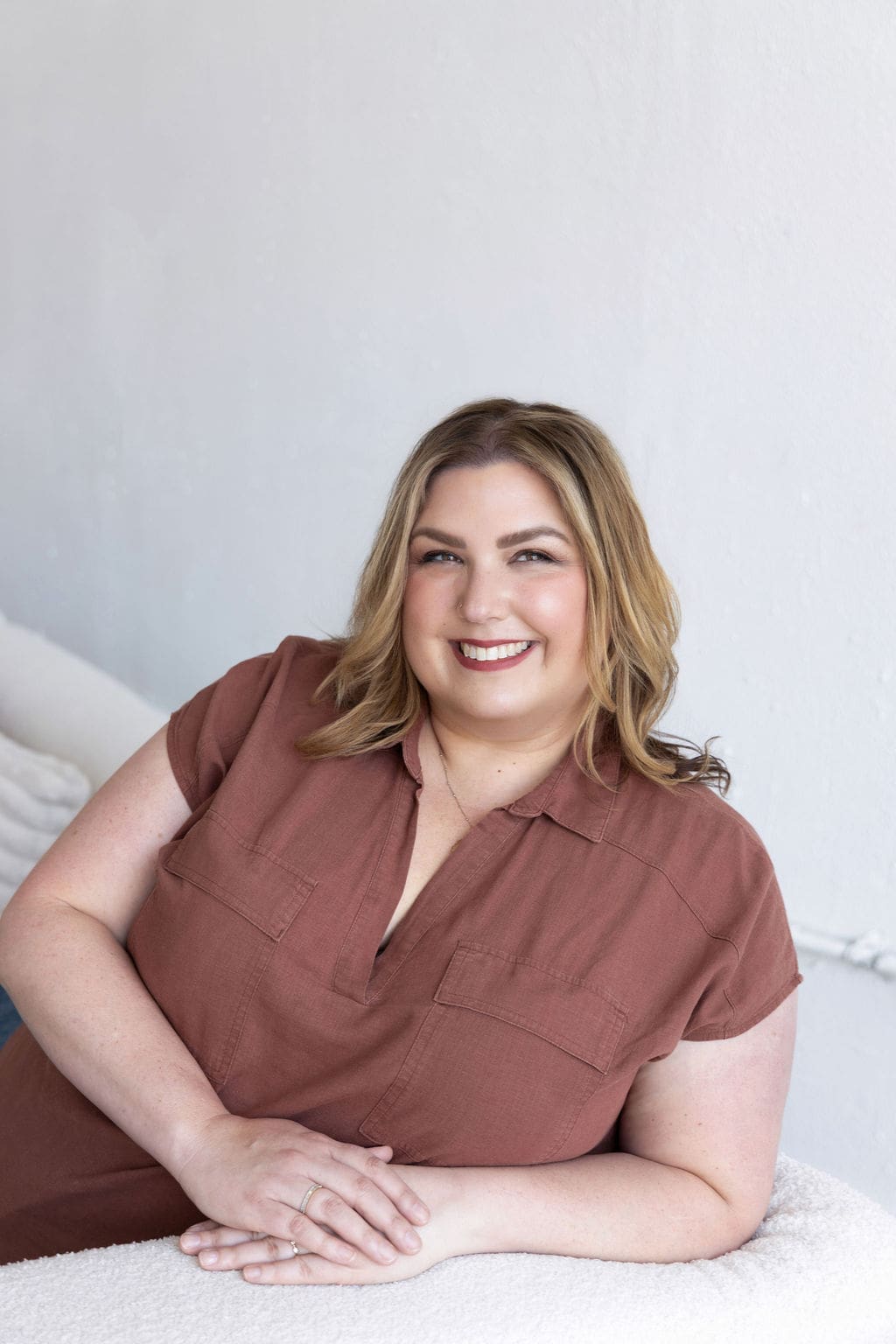The Empowered Therapist wearing a brown shirt and sitting on a white couch, smiling off into the distance with her hand on her face.