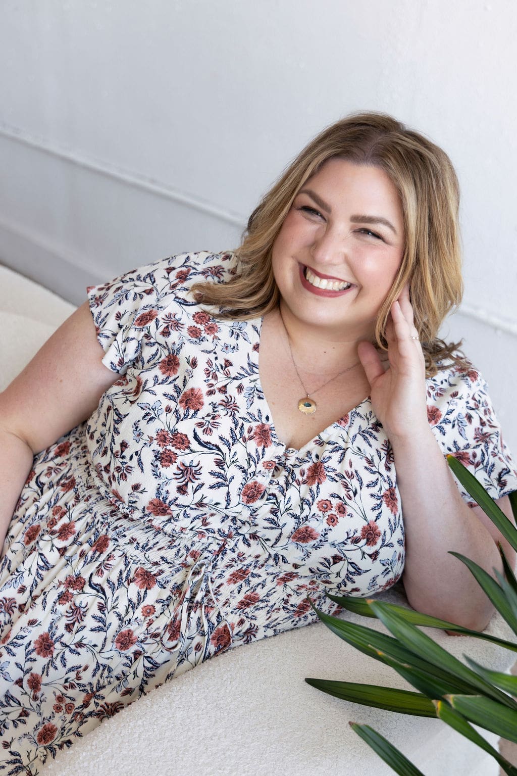 The empowered therapist and therapist mentor sitting on a white couch in a floral dress, smiling at the camera