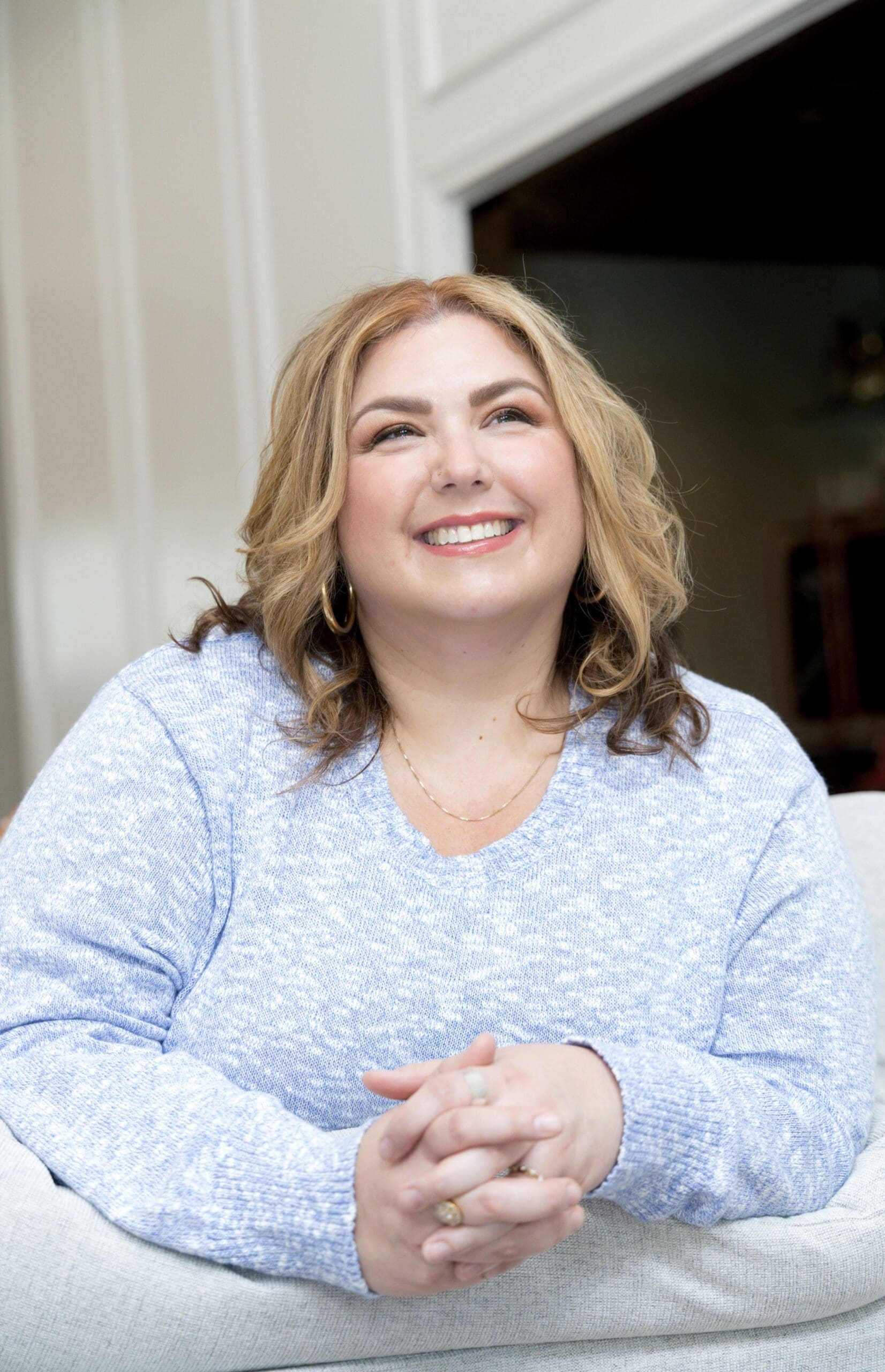 The Empowered Therapist, somatic experiencing practitioner, and therapist mentor, wearing a blue and white sweater leaning over a white couch, clasping her hands in front of her body and smiling into the distance.
