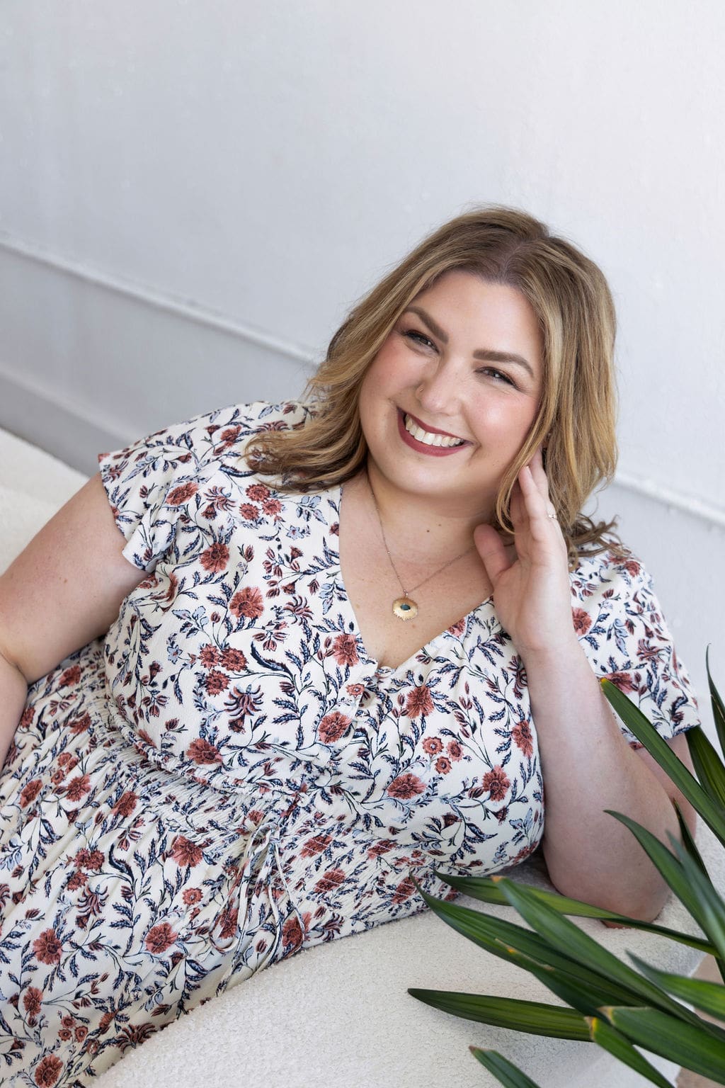 The Empowered Therapist and somatic experiencing practitioner sitting on a white couch in a floral dress, with her hand on her face smiling at the camera.