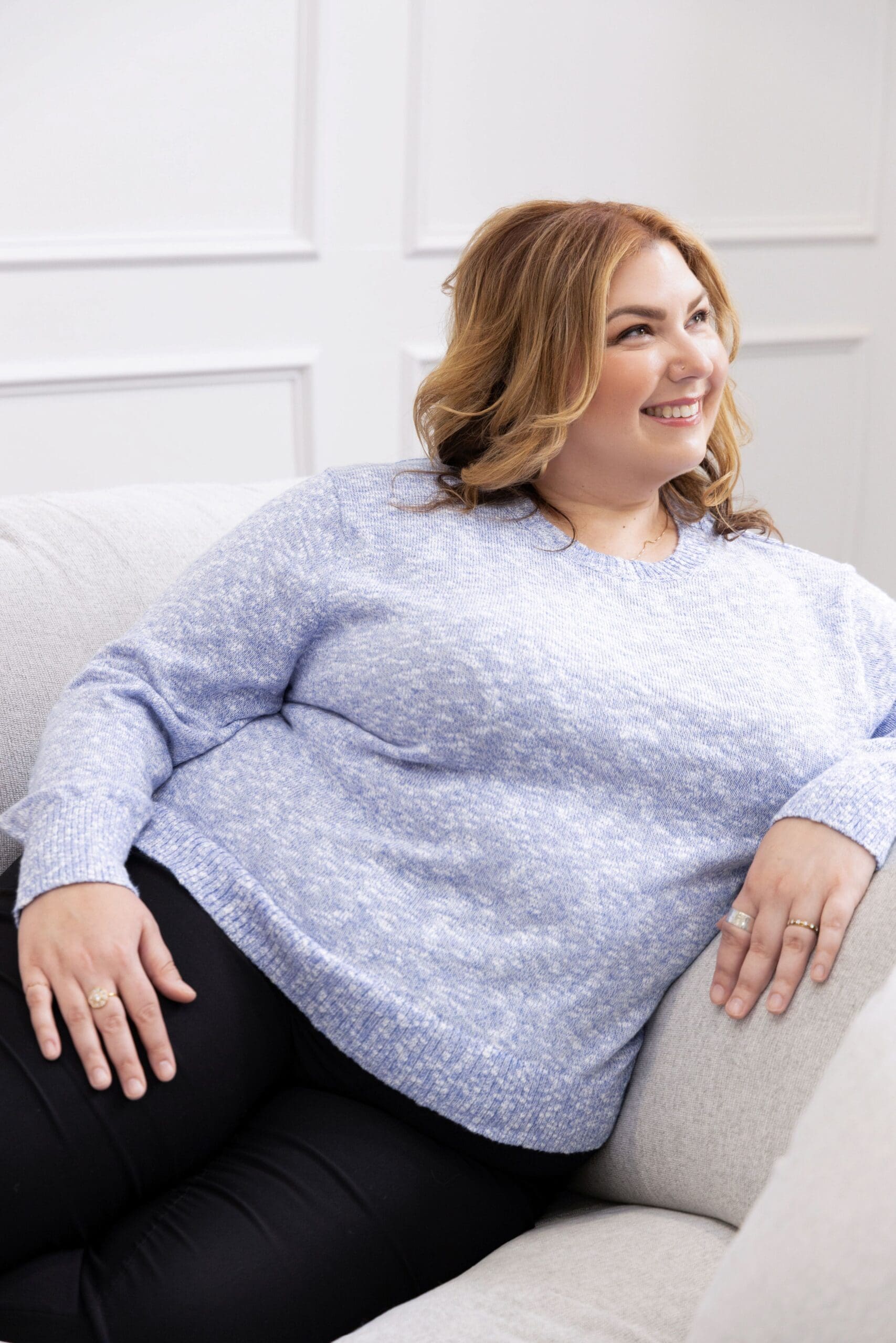 Therapist mentor and somatic experiencing practitioner wearing a blue sweater, lounging on a white couch and smiling off into the distance.
