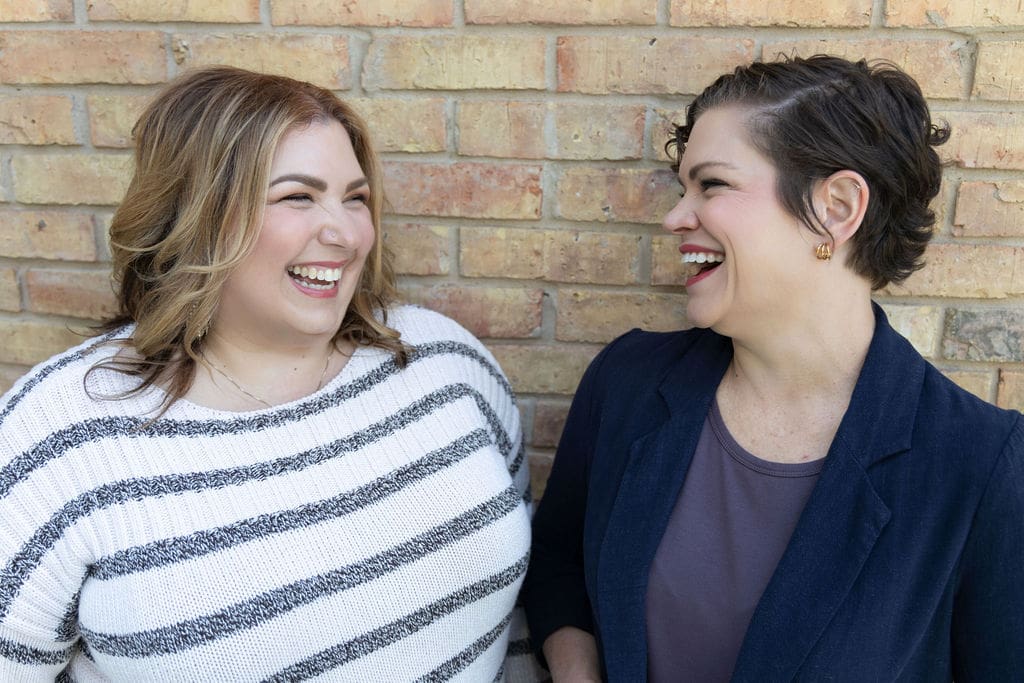 The Empowered Therapist and therapist mentor wearing a striped sweater standing in front of a brick wall, smiling and laughing in conversation with another somatic experiencing practitioner.