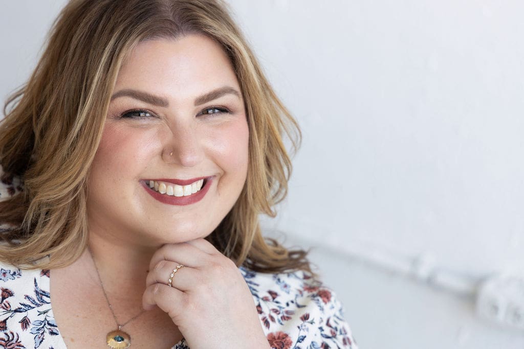 The empowered therapist and somatic experiencing practitioner wearing a floral top on a white background, with her hand under her chin, smiling at the camera.