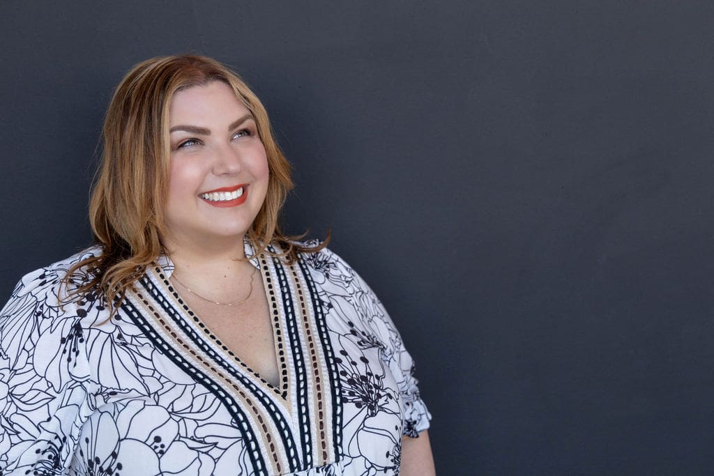 The empowered therapist and therapist mentor wearing a black and white floral patterned shirt in front of a black background, smiling into the distance.