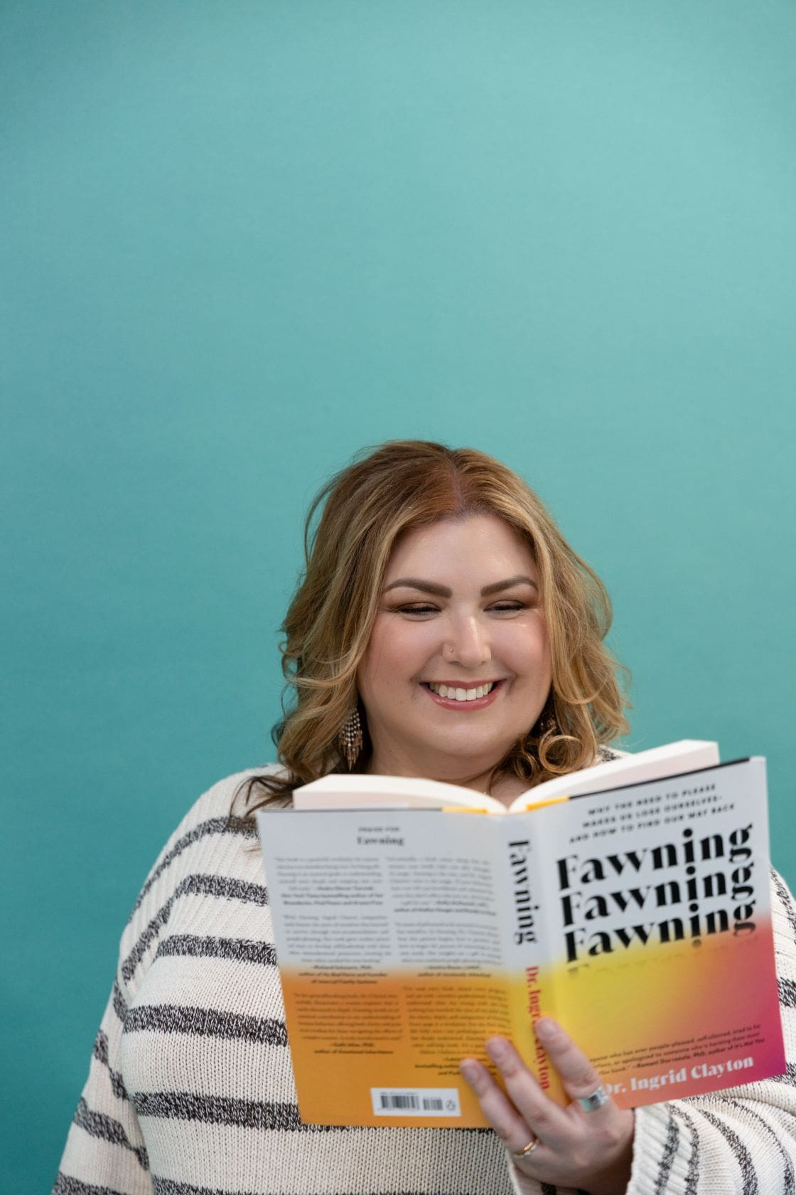 The empowered therapist and somatic experiencing practitioner wearing a striped sweater in front of a blue background, with hands over her heart and smiling into the distance.